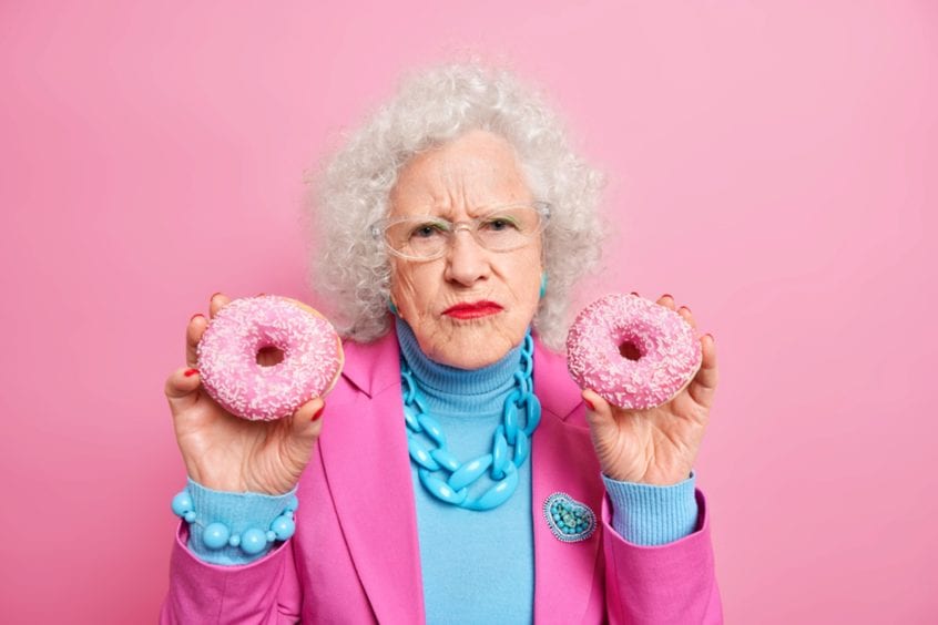 photo of an elderly woman wearing bright blue and pink holding two pink donuts, looking concerned