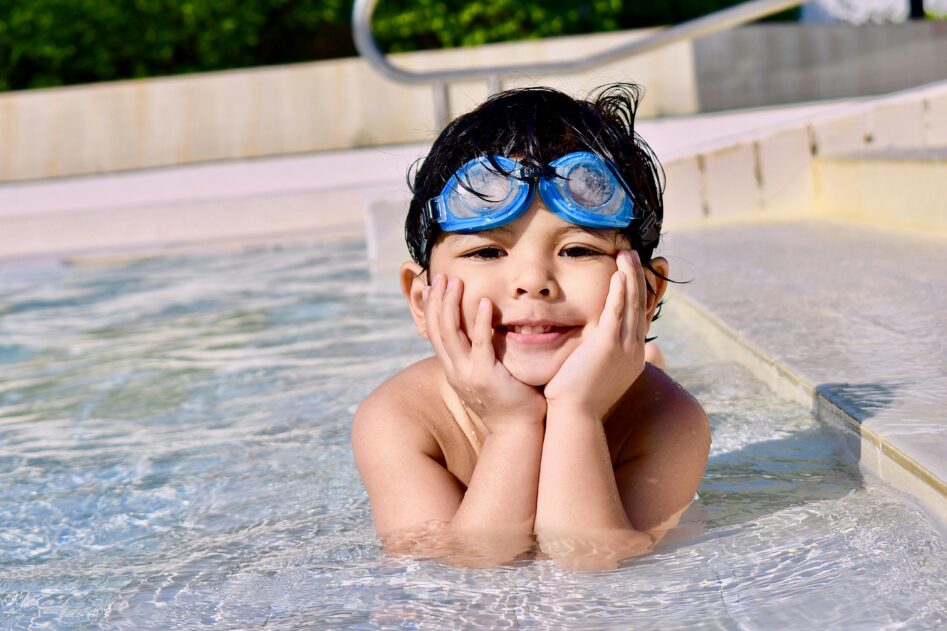 little boy laying in the shallow end of a pool with his chin resting on his hands, he is looking straight into the camera. He has dark brown hair and blue goggles on his head.